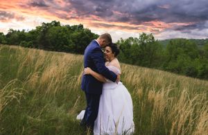 Bride and groom embracing in golden field at sunset with dramatic purple sky backdrop.