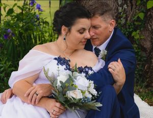 Bride and groom share a tender moment under a tree, bride holding a white and blue bouquet in a white off-shoulder gown.
