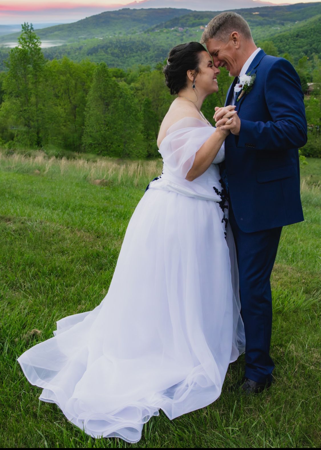 A couple shares a romantic dance in a white wedding gown and blue suit on a lush green hillside at sunset.
