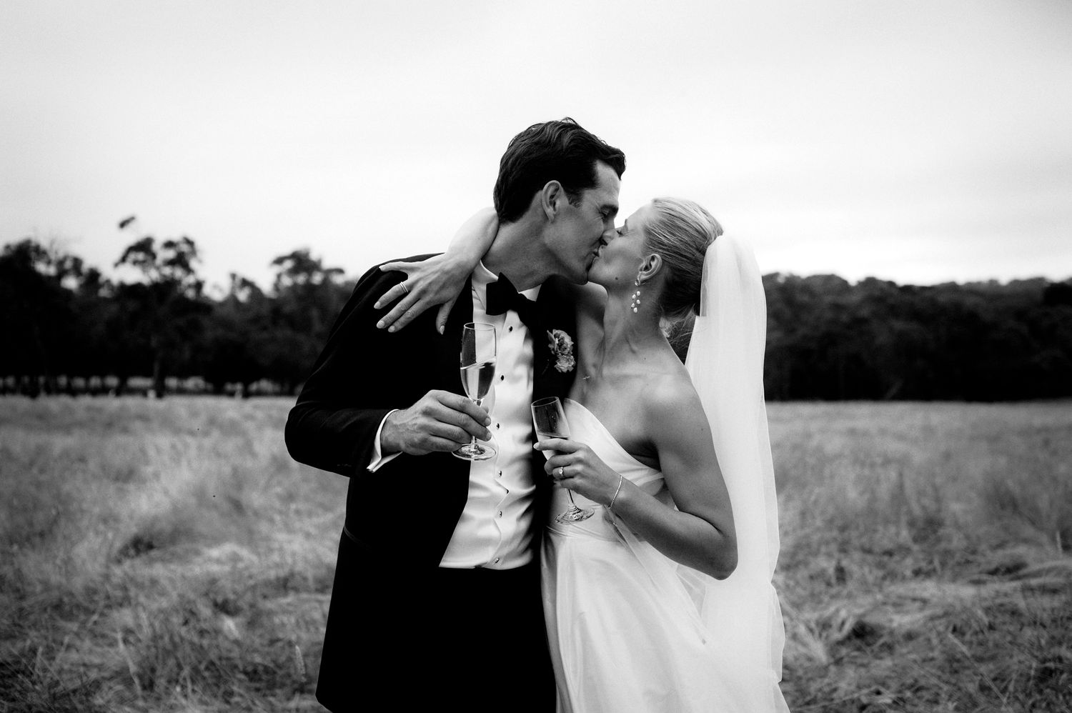Black and white photo of a bride and groom sharing a kiss in a field, holding champagne glasses on their wedding day.