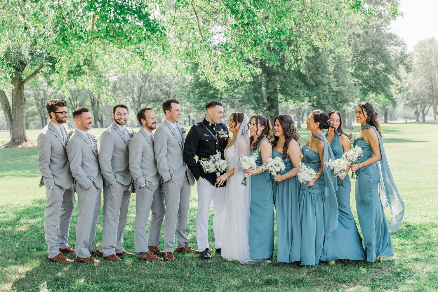 Wedding party in gray suits and blue dresses posing outdoors under green trees on a sunny day.