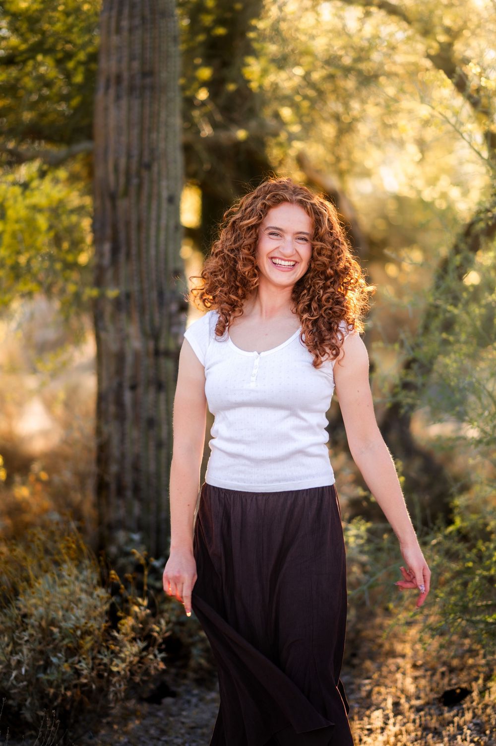 Young woman with curly red hair smiles outdoors in white top and black skirt, surrounded by lush green trees.