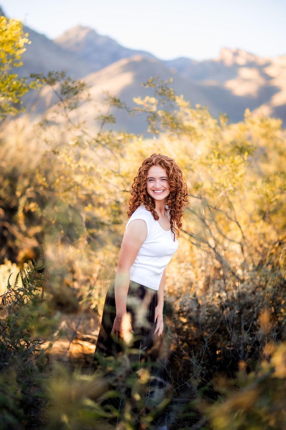 Smiling young woman with curly red hair in white top stands among golden desert shrubs with mountains behind her.