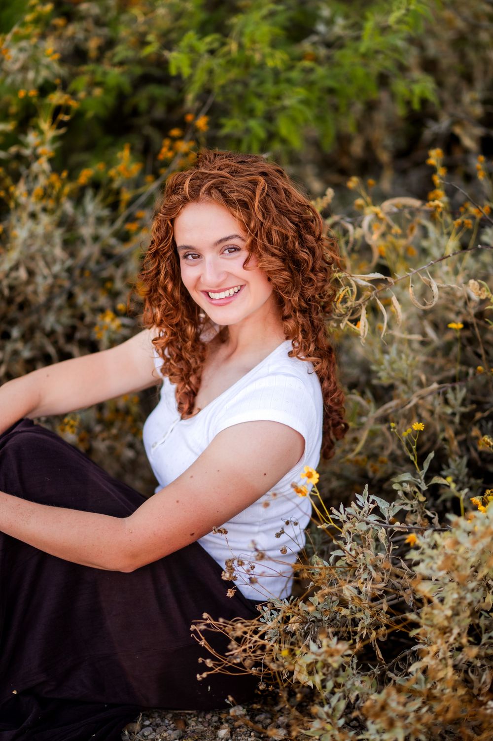 Young woman with curly red hair smiles outdoors, sitting among wildflowers and green shrubs in natural light.