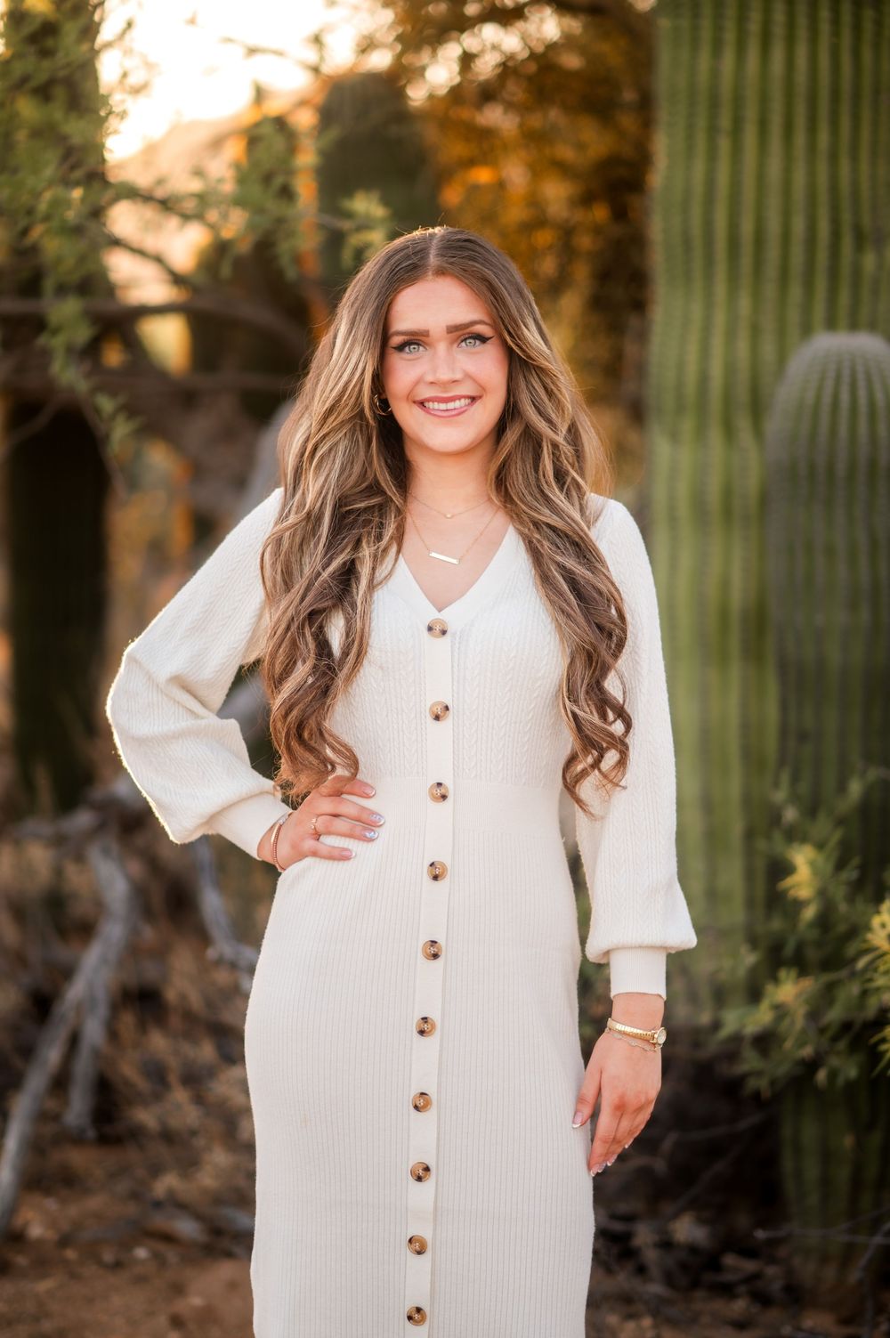 Young woman in cream button-down midi dress smiling outdoors near saguaro cactus with long wavy hair.