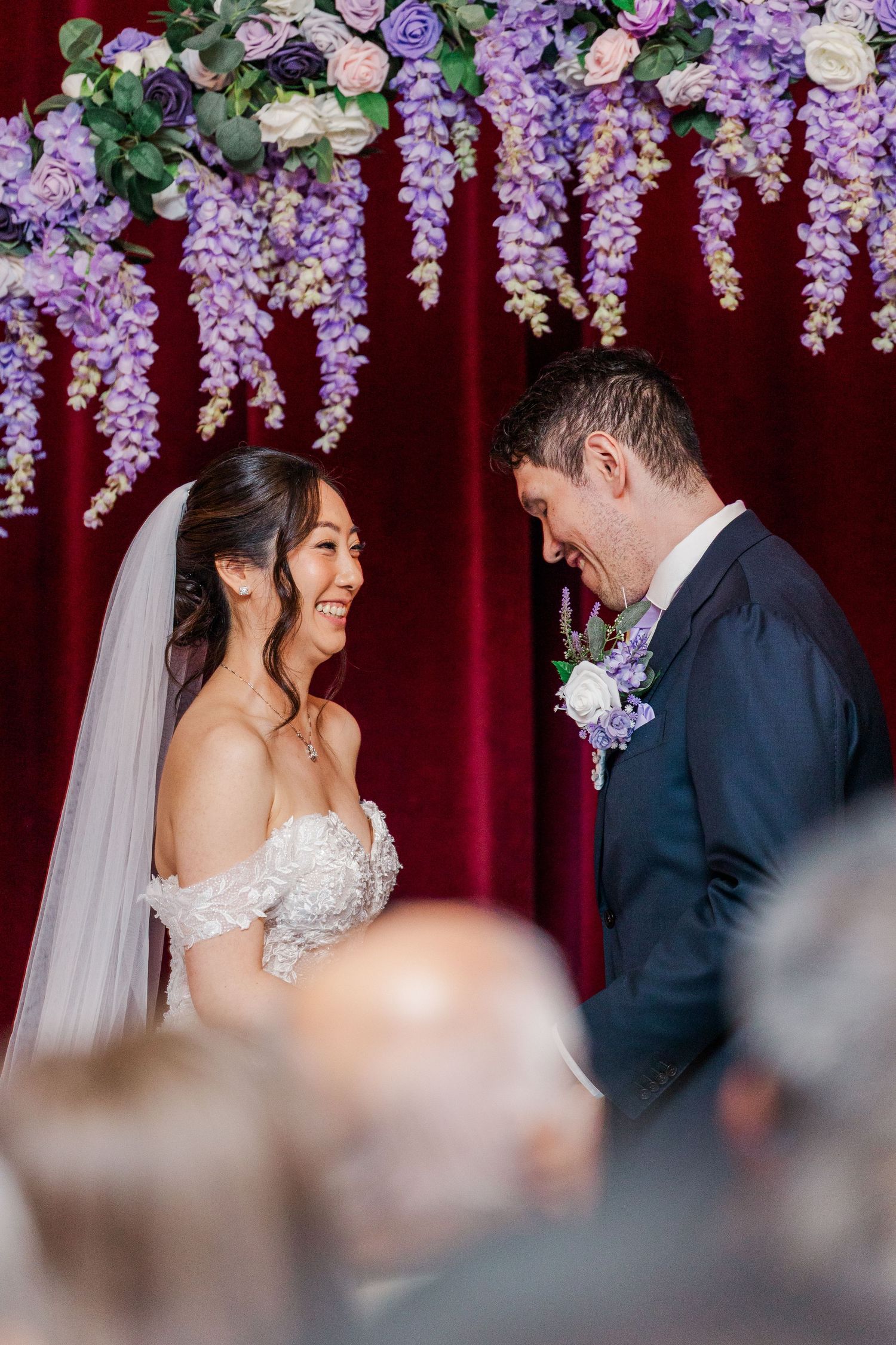 Bride and groom share a joyful moment at altar adorned with purple wisteria and roses against red velvet backdrop.