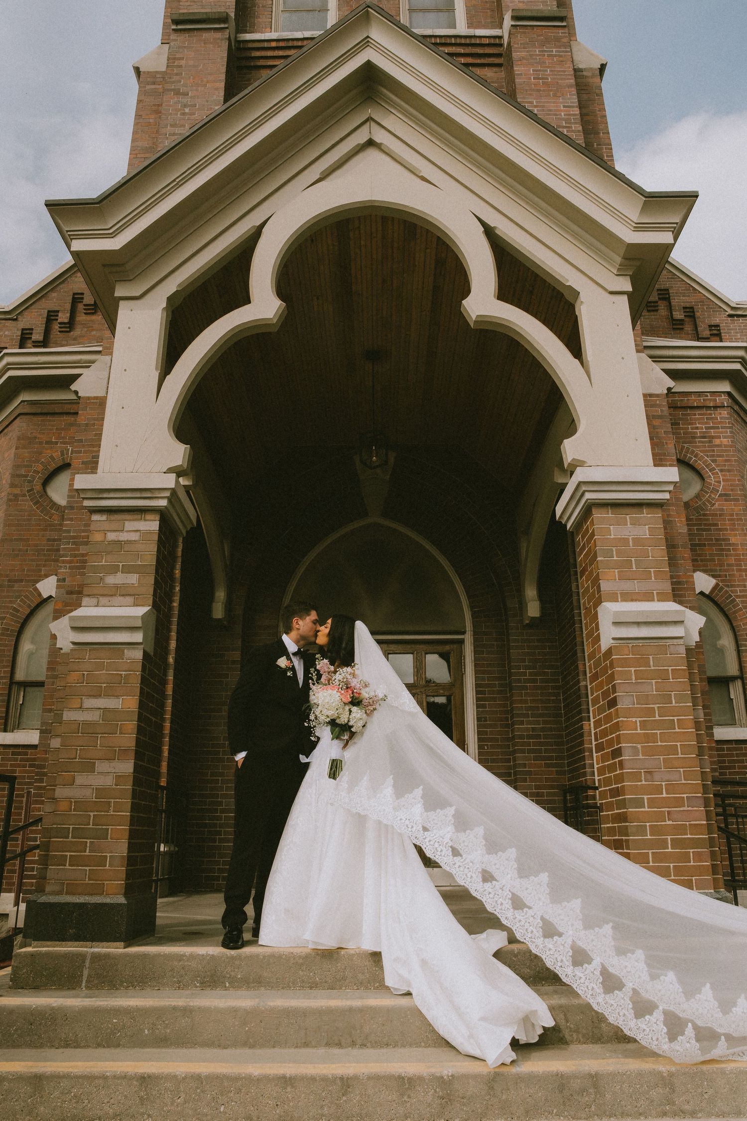 Bride and groom kiss on church steps, her lace veil flowing in the wind before a classic brick building.