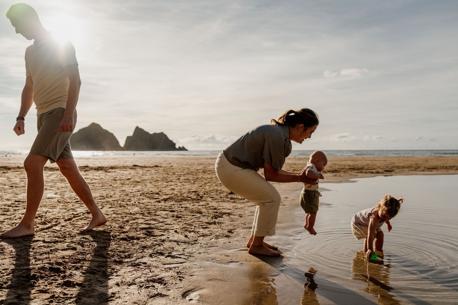 Family enjoying a sunny beach day, with parents watching toddlers play in shallow ocean water on sandy shore.