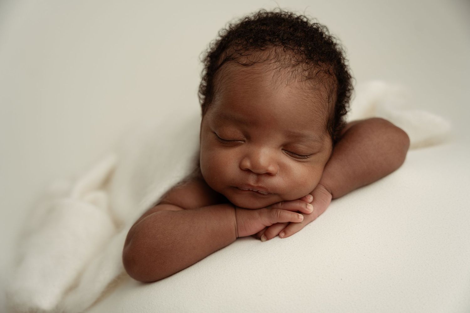 Peaceful newborn baby sleeping with hands folded under chin on white blanket in professional studio portrait.