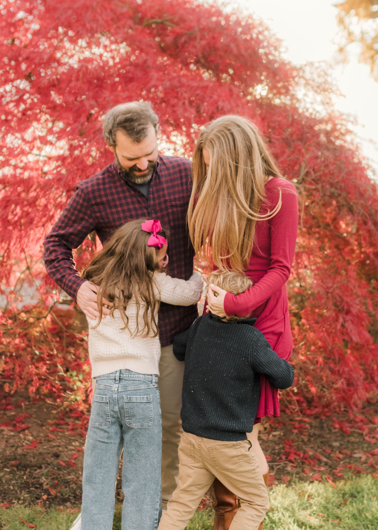Family of four embracing outdoors in autumn, surrounded by vibrant red foliage during a fall photo session.
