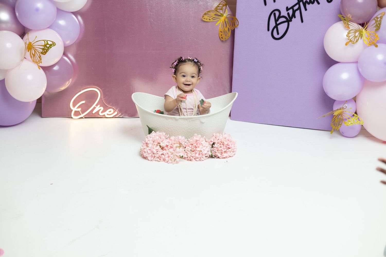 Baby girl sits in a mini bathtub at a purple balloon first birthday cake smash photo shoot.