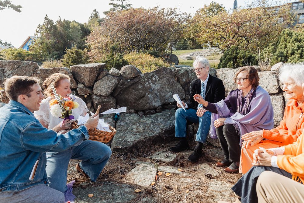 Group of people sitting outdoors on rocks, engaged in conversation surrounded by autumn foliage and stone walls.