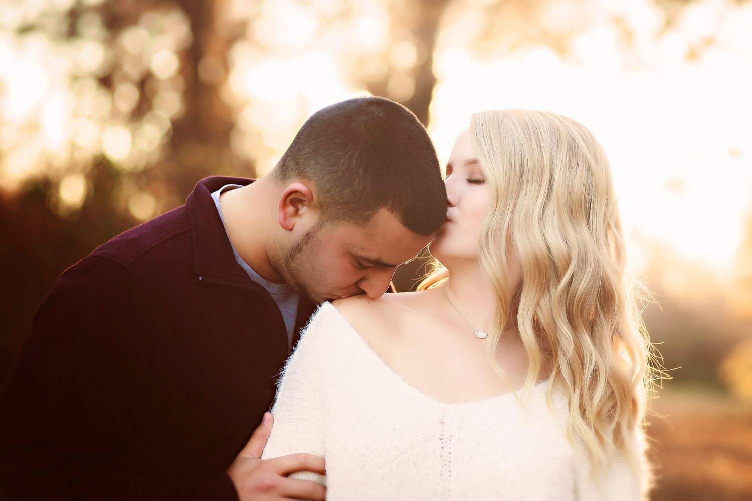 Romantic couple sharing a tender moment outdoors in warm golden light, woman in white sweater kissing man's head.