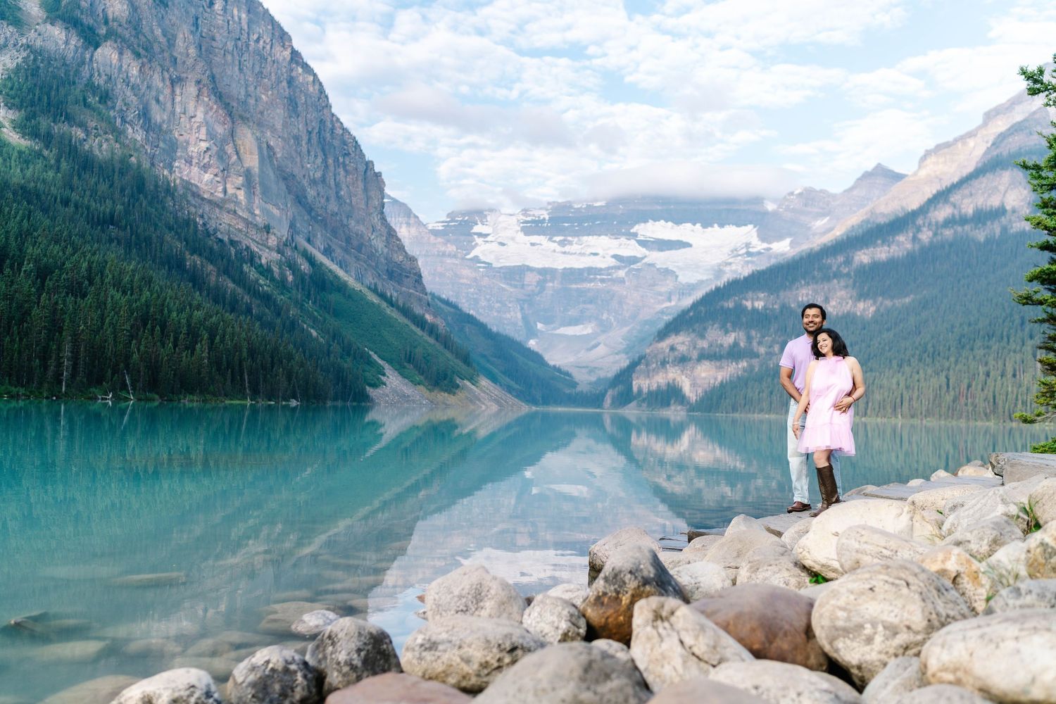 Couple standing on rocky shore of turquoise Lake Louise with stunning mountain reflections in Banff National Park, Canada.