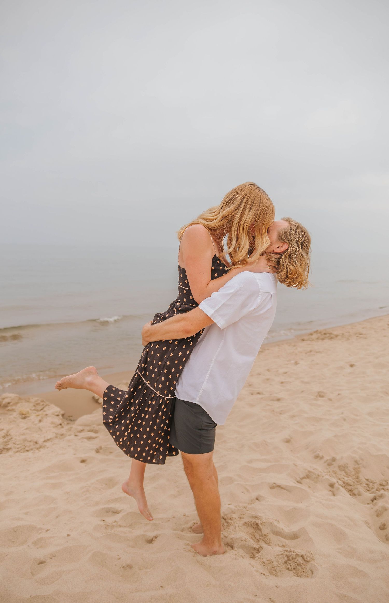 A couple shares a romantic kiss on a sandy beach, as the man lifts the woman in a flowing floral dress.