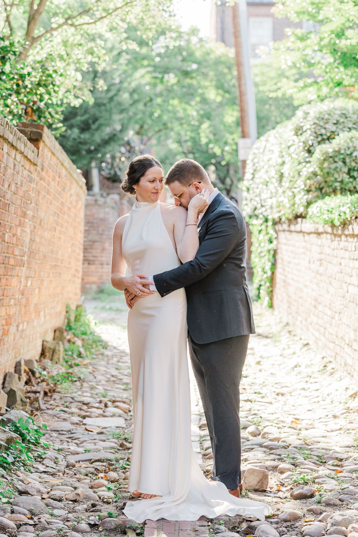 Bride and groom share a tender moment on a cobblestone alley, framed by brick walls and lush greenery.