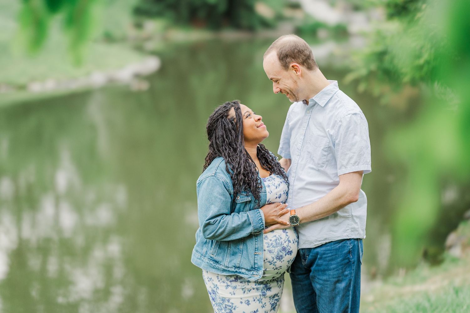 Interracial couple sharing a tender moment outdoors near a pond, woman in floral dress and denim jacket, man in blue shirt.
