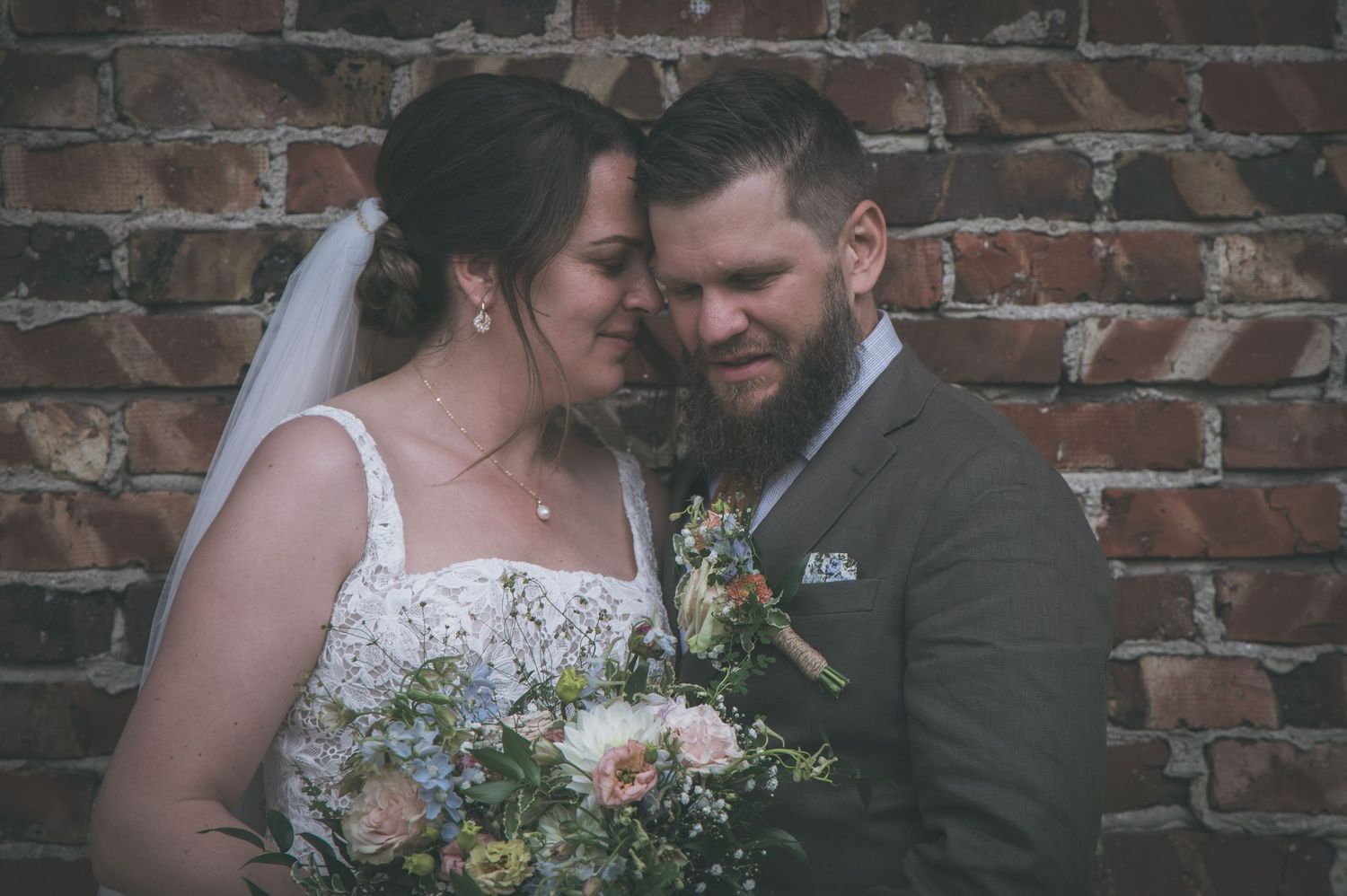 Romantic couple embraces tenderly against rustic brick wall, bride holding pastel wildflower bouquet.