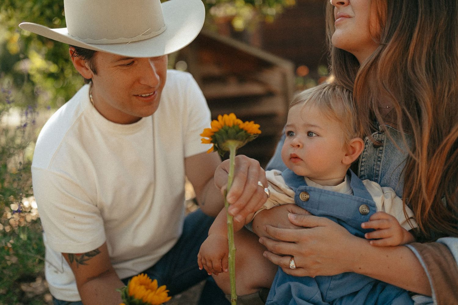 A man in a white tee and cowboy hat holds a sunflower near a baby in denim overalls held by a woman outdoors.