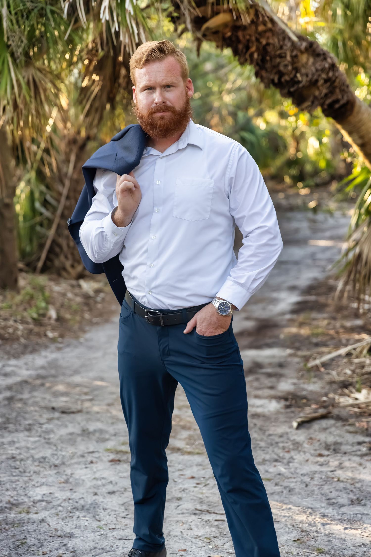 Red-bearded man in white shirt and navy pants holds jacket over shoulder on a sandy tropical path.