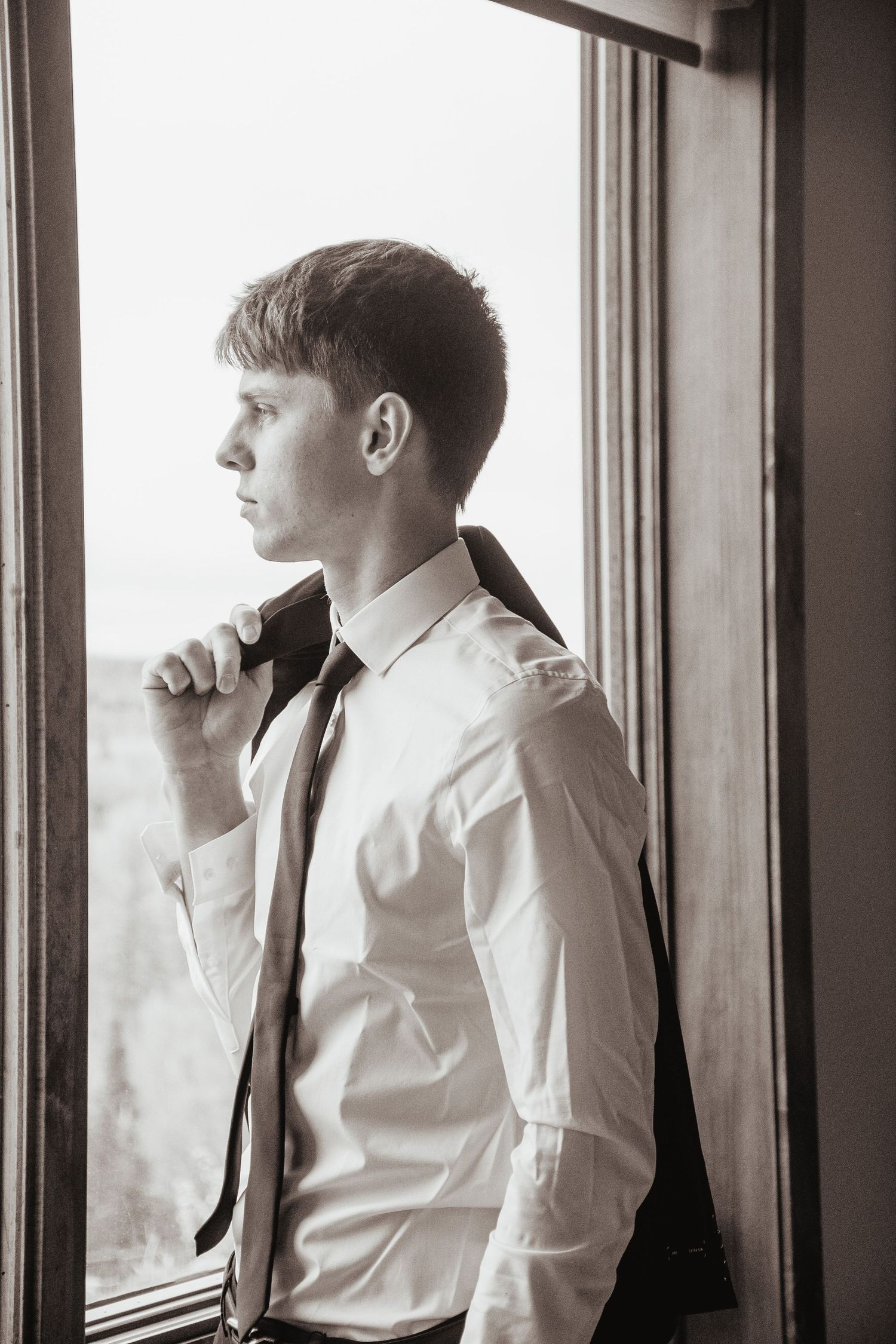 Young man in white dress shirt and tie holds jacket over shoulder, gazing thoughtfully out a window in black and white photo.