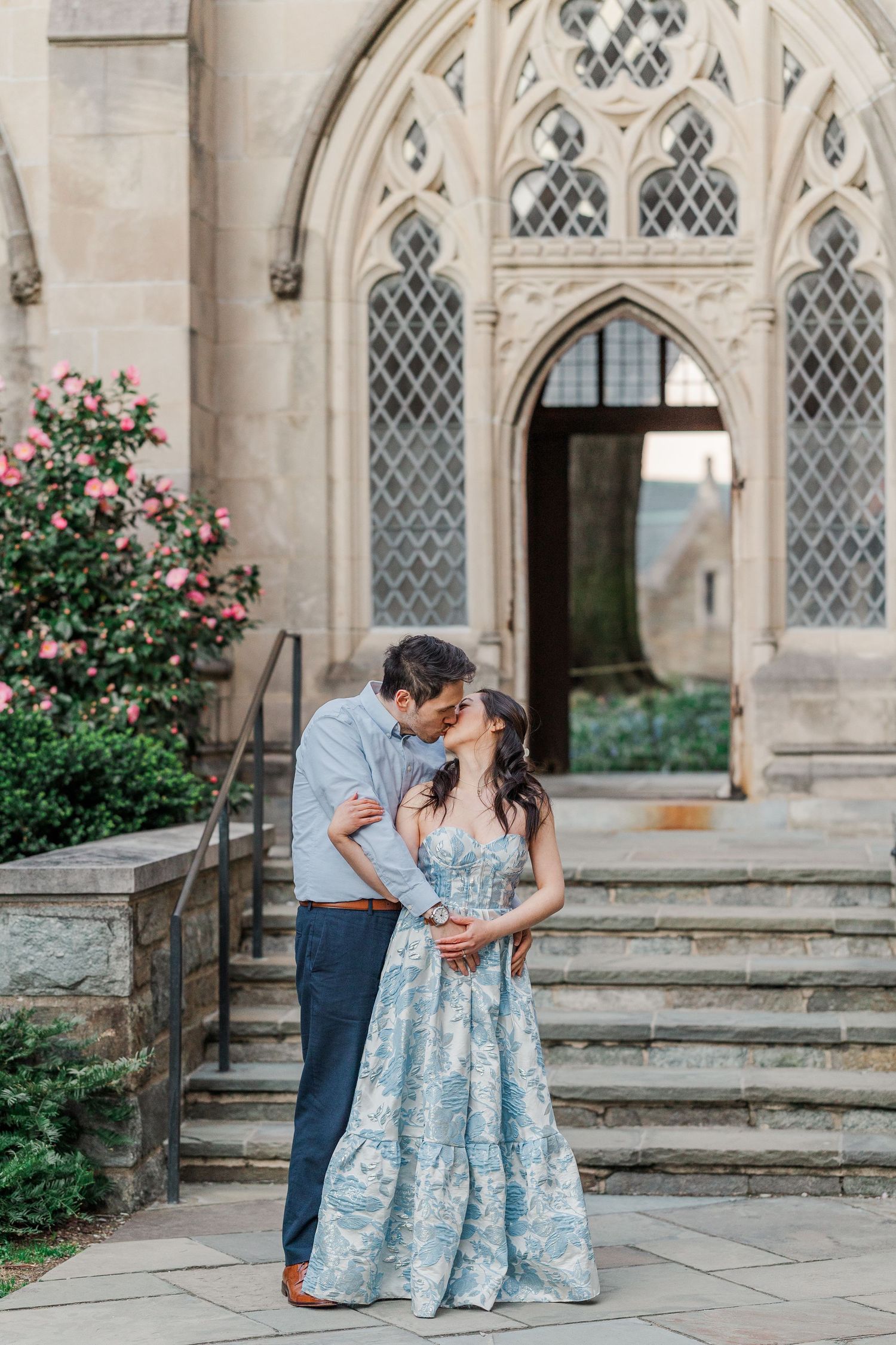 Couple sharing a romantic kiss in front of a Gothic stone archway, woman in blue floral maxi dress.