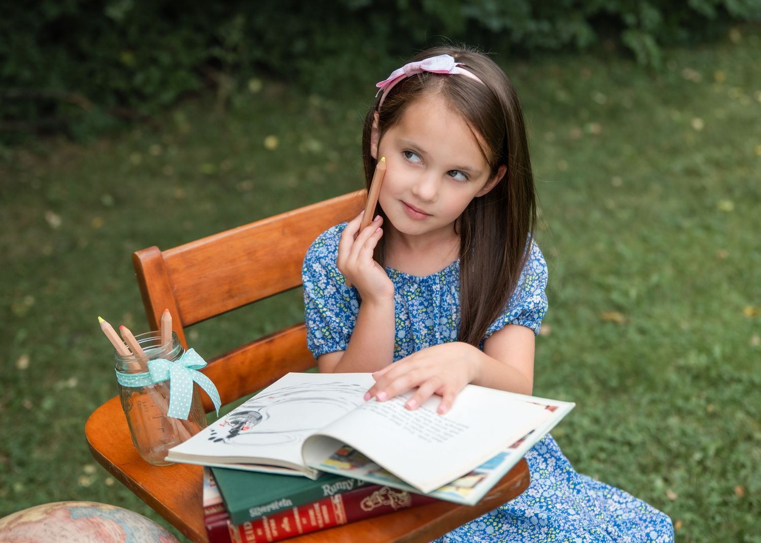 Young girl in blue dress reading a book outdoors on a wooden chair, surrounded by stacked books and pencils.