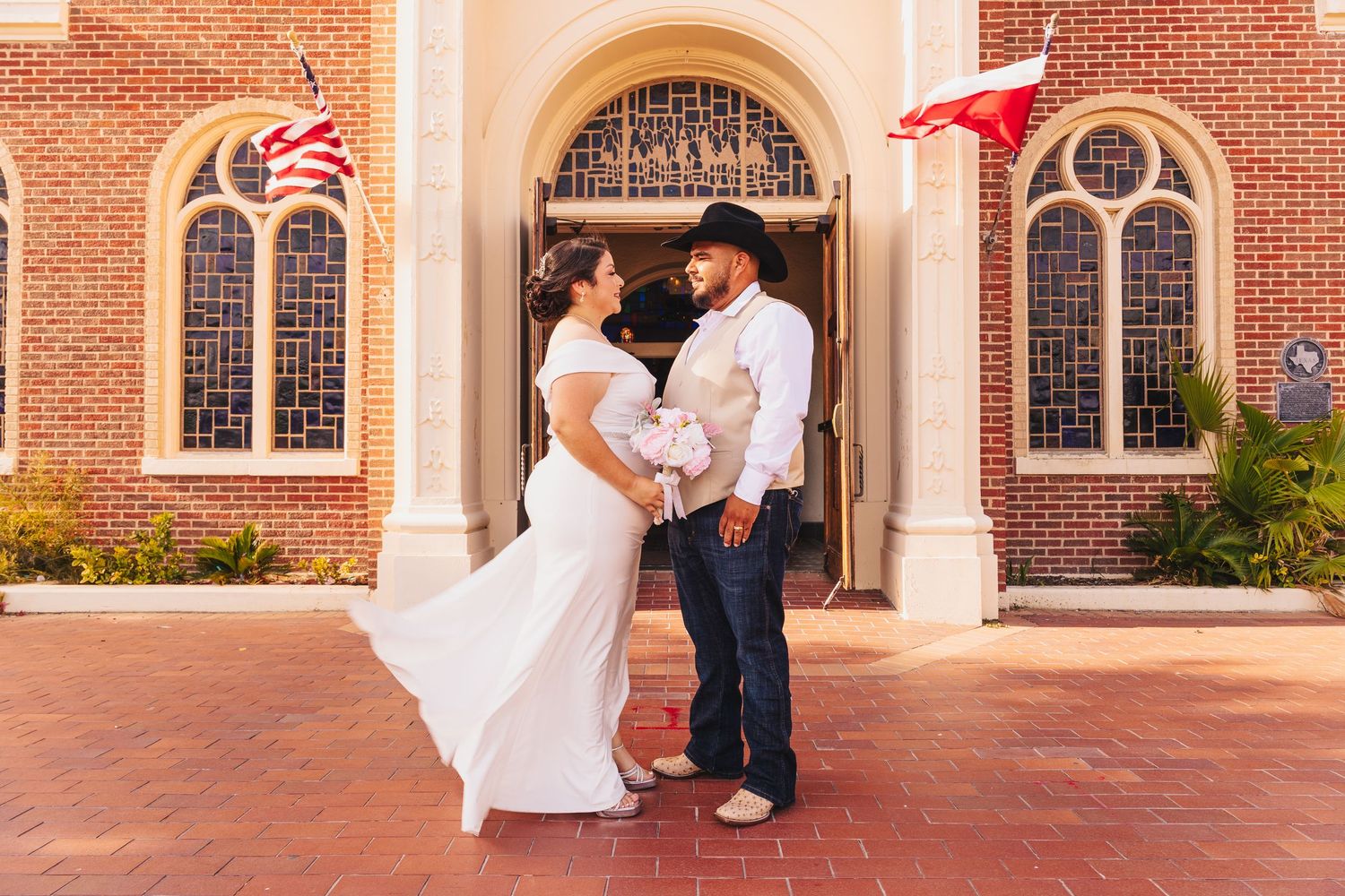 Bride and groom in cowboy hat stand facing each other outside a brick church with Texas and American flags.
