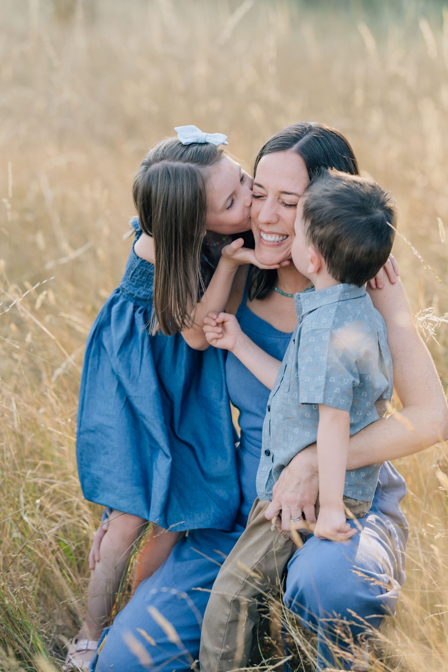 A smiling mother sits in a golden field as her young daughter and son kiss her cheeks outdoors.