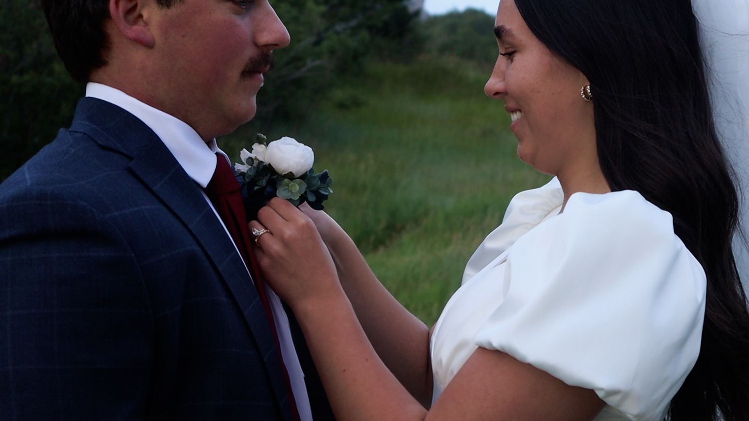 Bride and groom share a romantic moment outdoors, smiling at each other with lush green trees in the background.