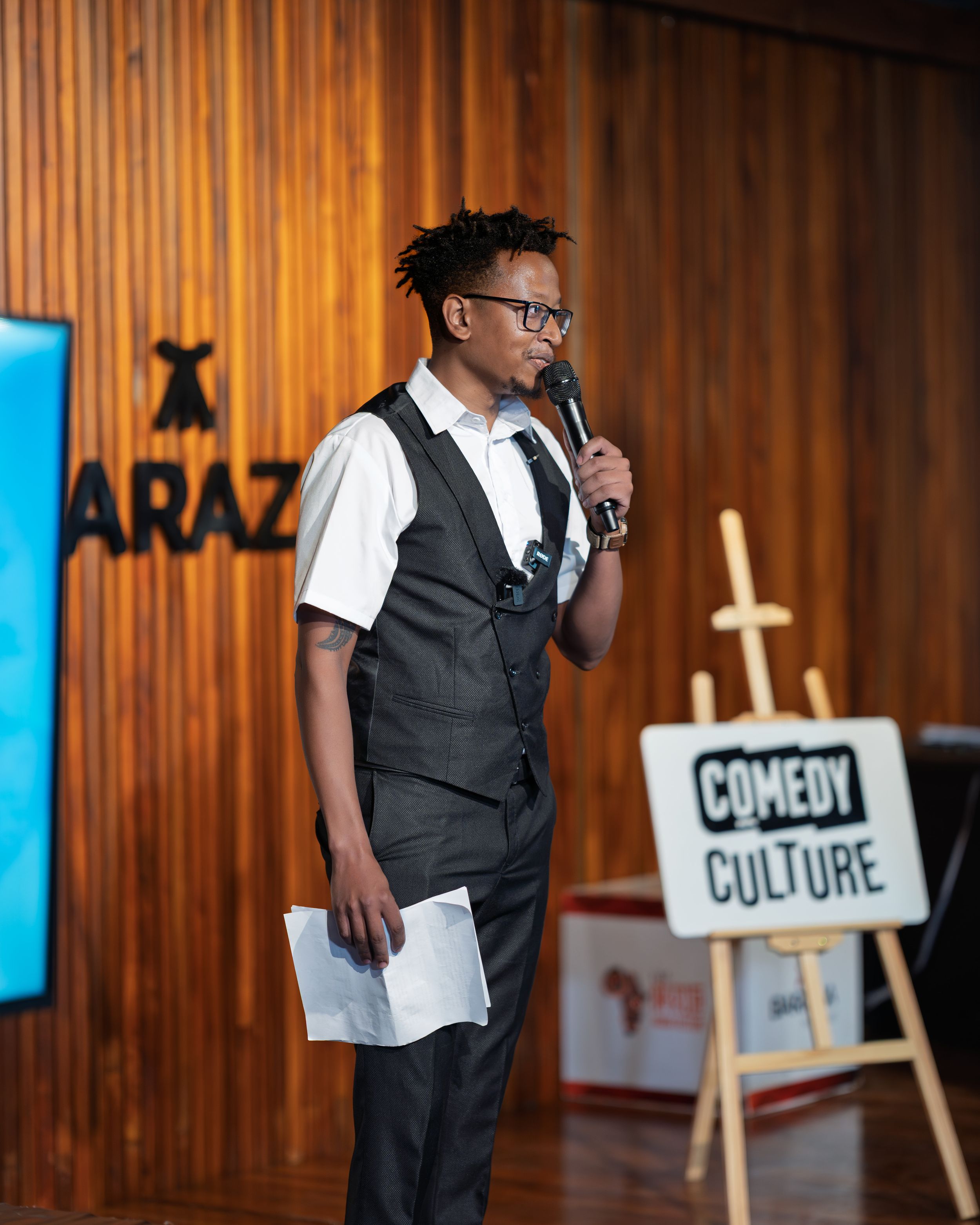 Young man in a vest and glasses hosting a Comedy Culture event, holding a microphone and papers.