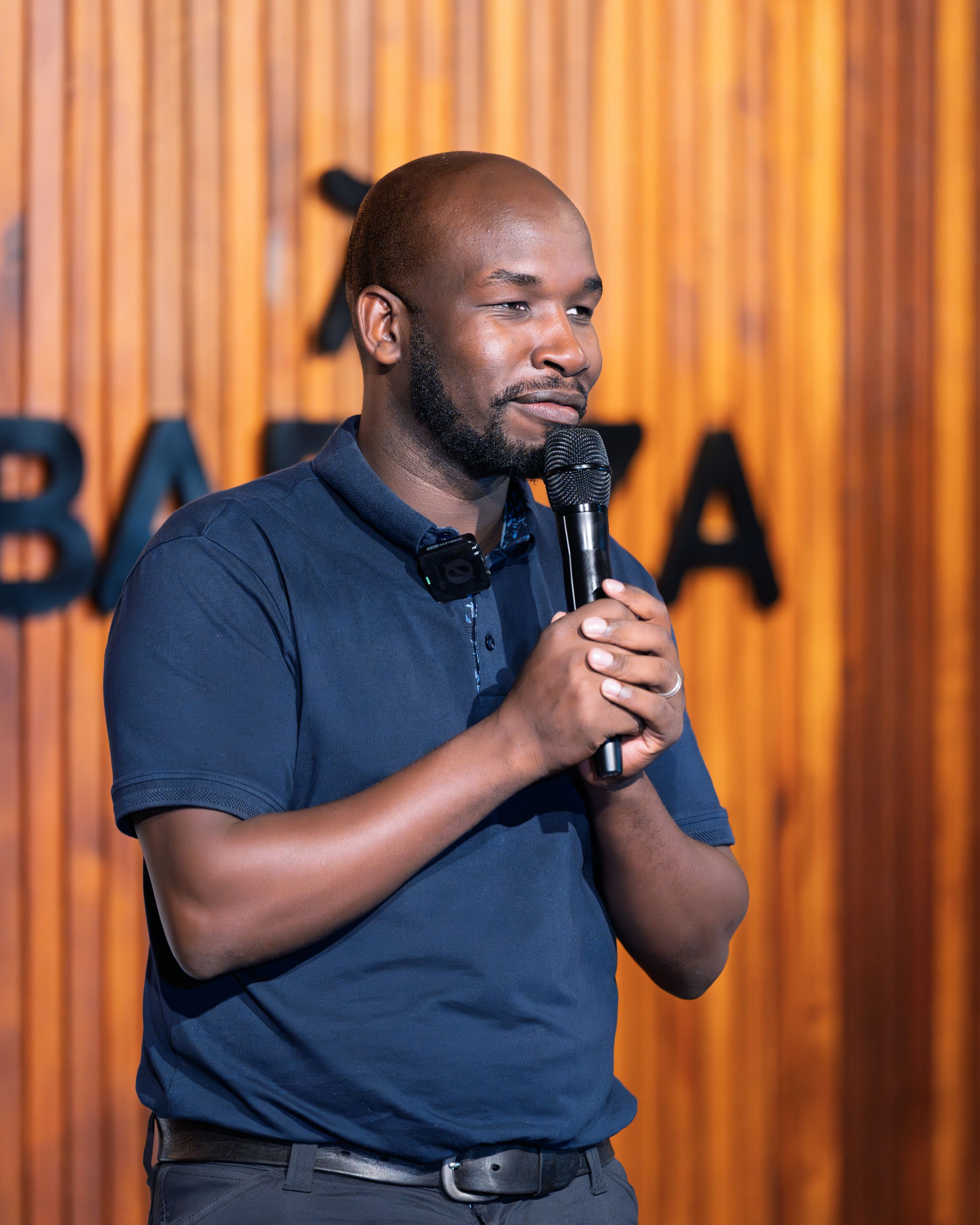 Man in navy polo shirt speaks into microphone at Barka event.