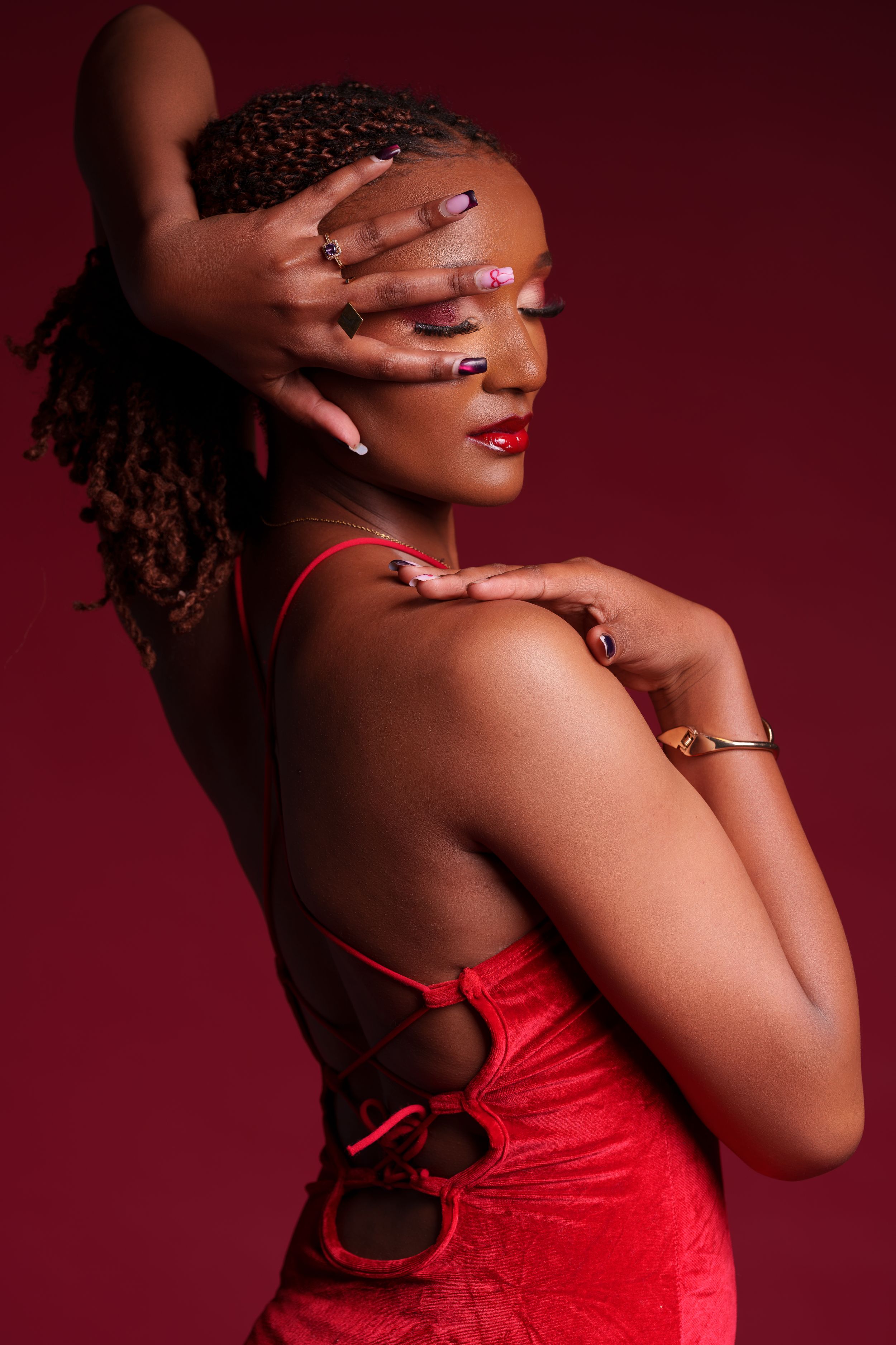 Young Black woman in red dress poses elegantly with hands covering face, showcasing long nails and rings against red background.