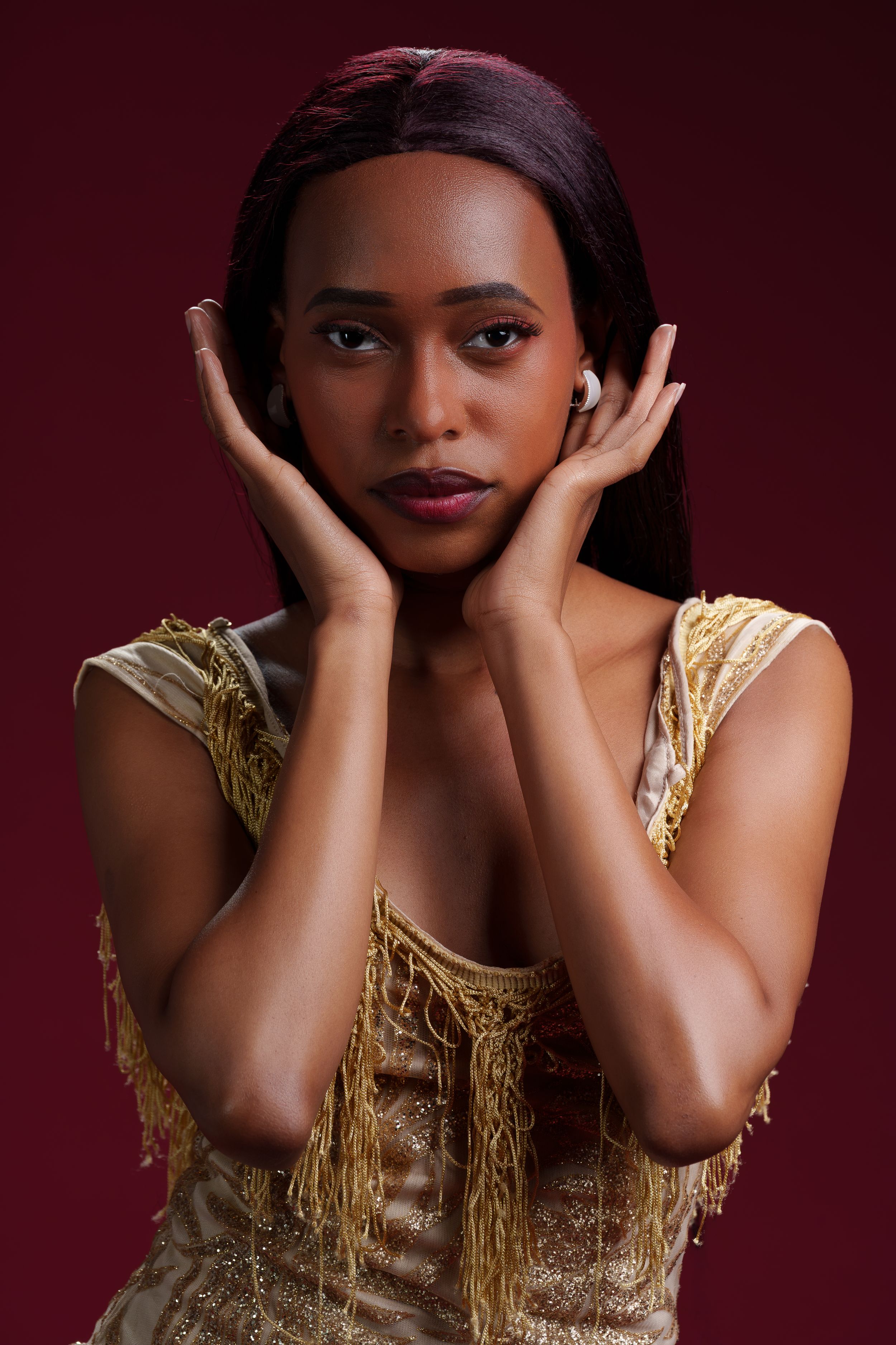 Elegant woman in gold dress poses with hands framing her face on red background.