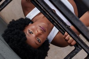 Young Black woman with natural afro hair looking up through metal bars, wearing a white top.