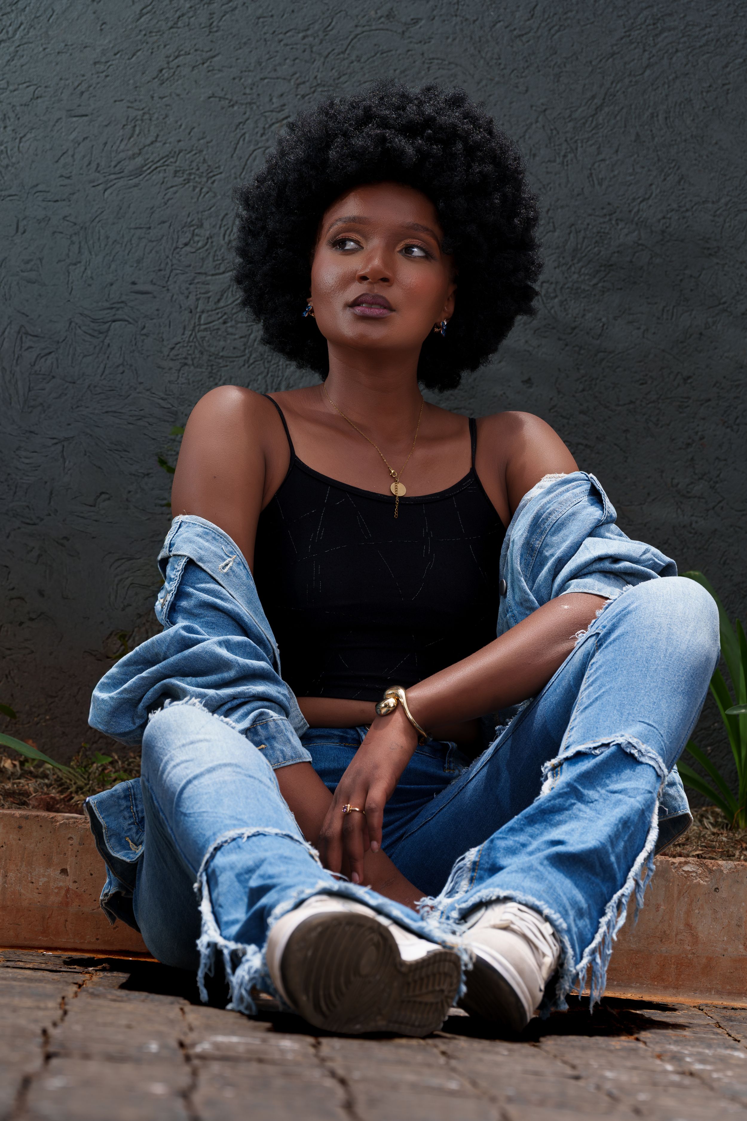 Young Black woman with afro hair sits cross-legged on ground wearing ripped jeans, black crop top, and denim jacket.