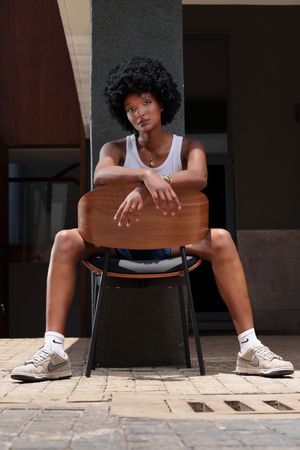 Young Black woman with afro hair sits on wooden chair outdoors, wearing white tank top, denim shorts, and Nike sneakers.