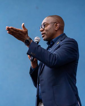Black man in navy blue suit speaks passionately into microphone with hand gesture against blue background.
