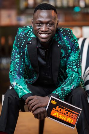 Smiling man in green patterned blazer holds Jokes Aside comedy show sign.