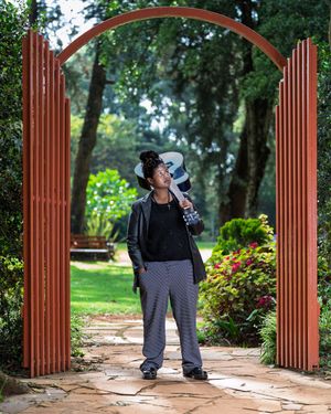 Woman holding a camera standing under a red archway in a lush green garden park setting.