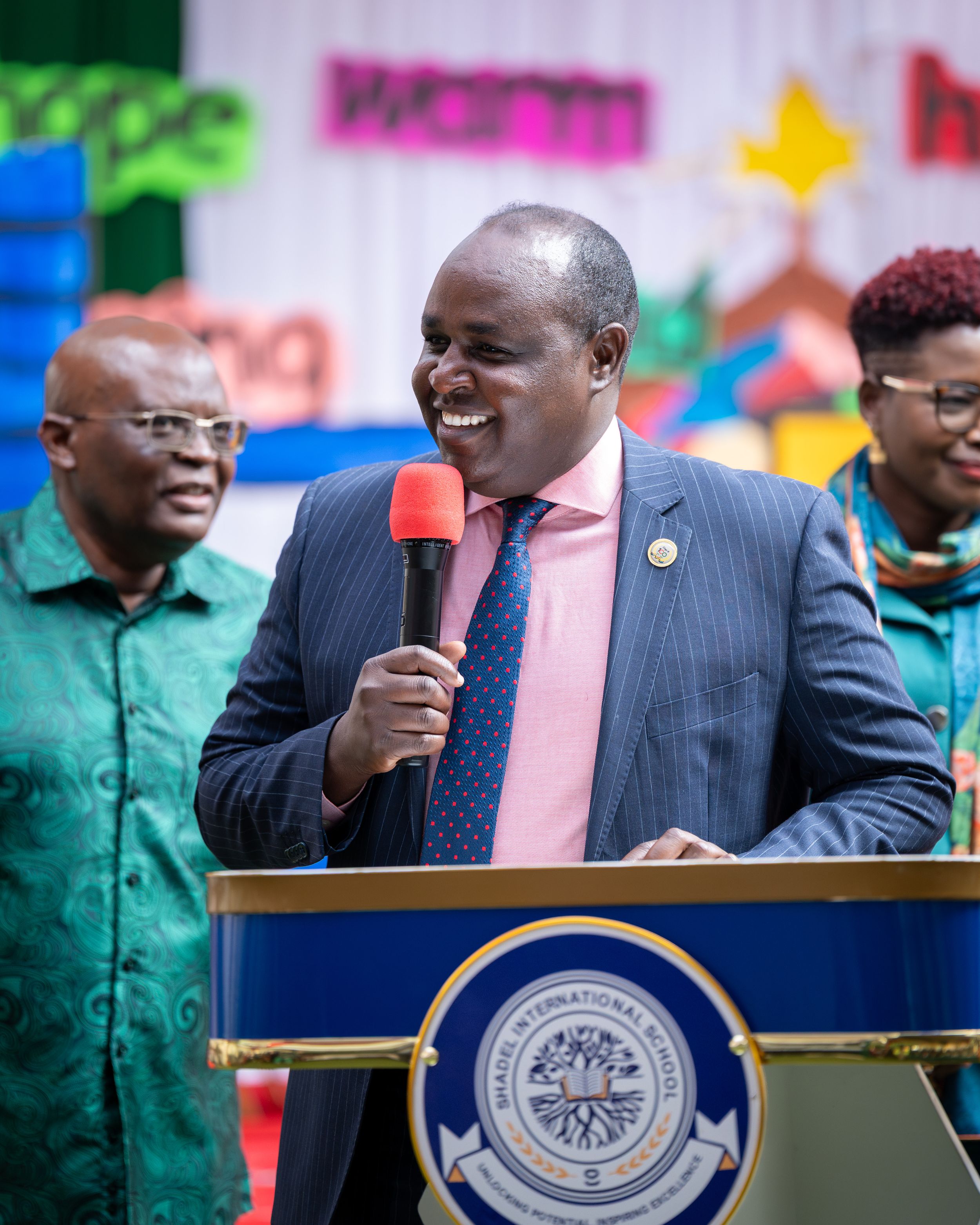 Smiling man in a blue pinstripe suit speaking into a red microphone at a podium during an event.