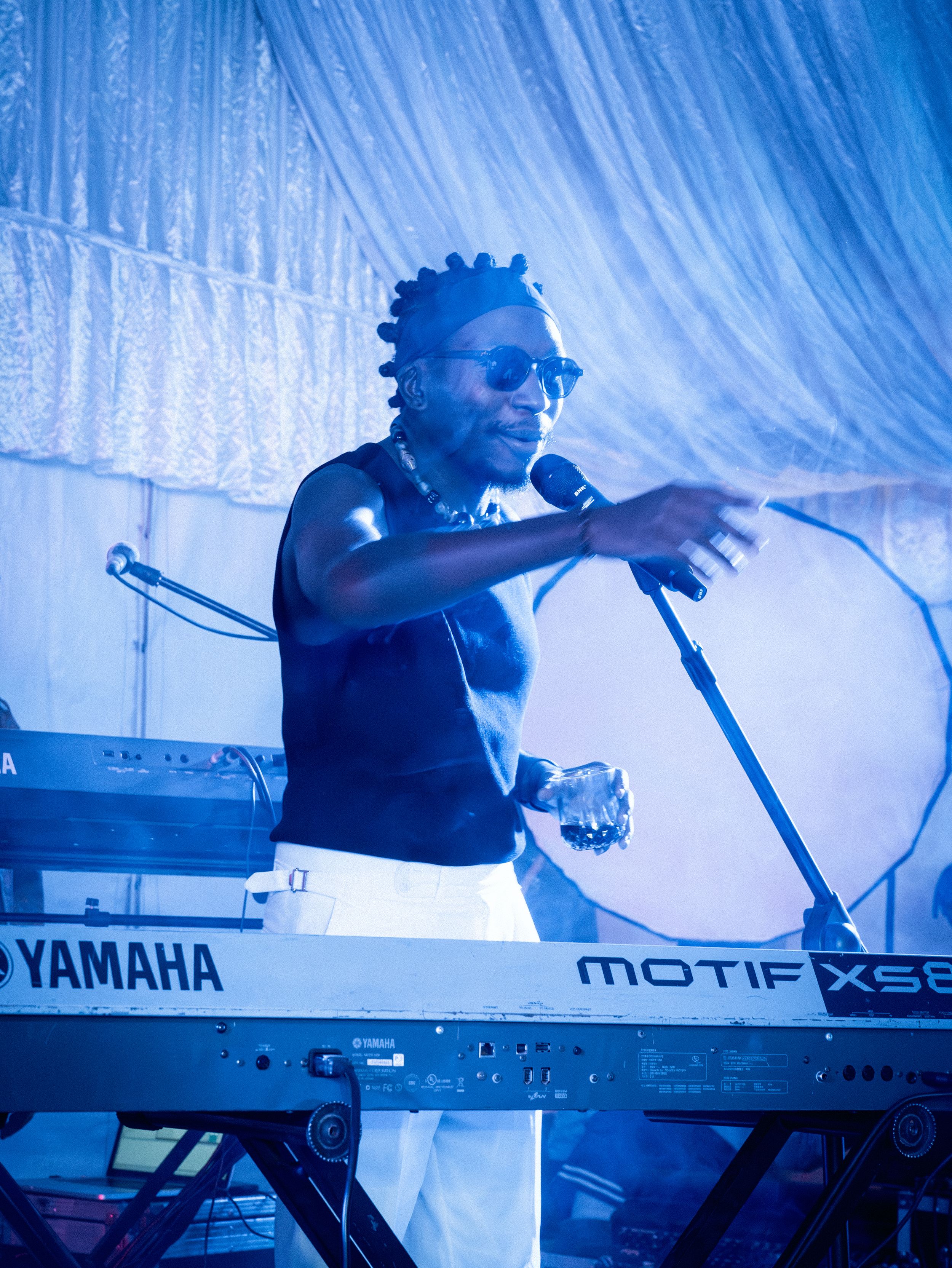 Musician playing a Yamaha Motif XS keyboard and singing into a microphone under blue stage lighting.