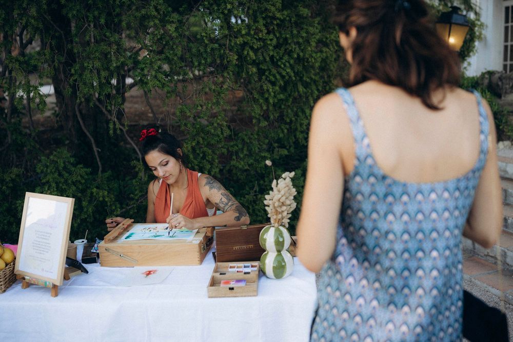 A tattooed woman creates calligraphy art at a white table outdoors while another woman in a blue dress watches.
