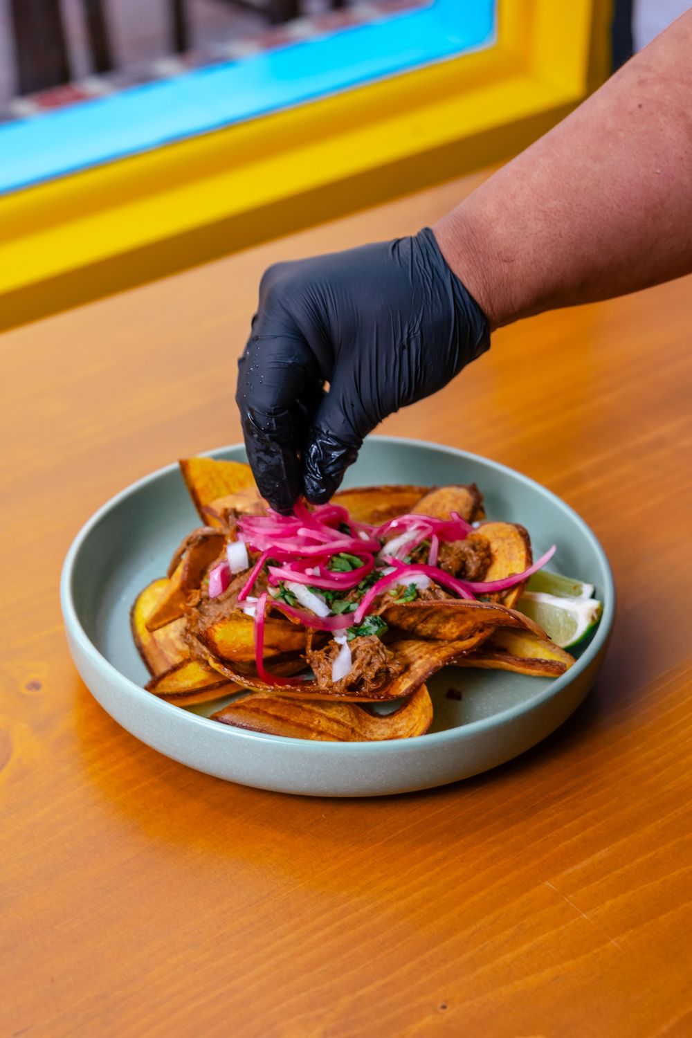 Chef garnishing a plate of crispy plantains with pickled red onions and pulled pork on a wooden table.
