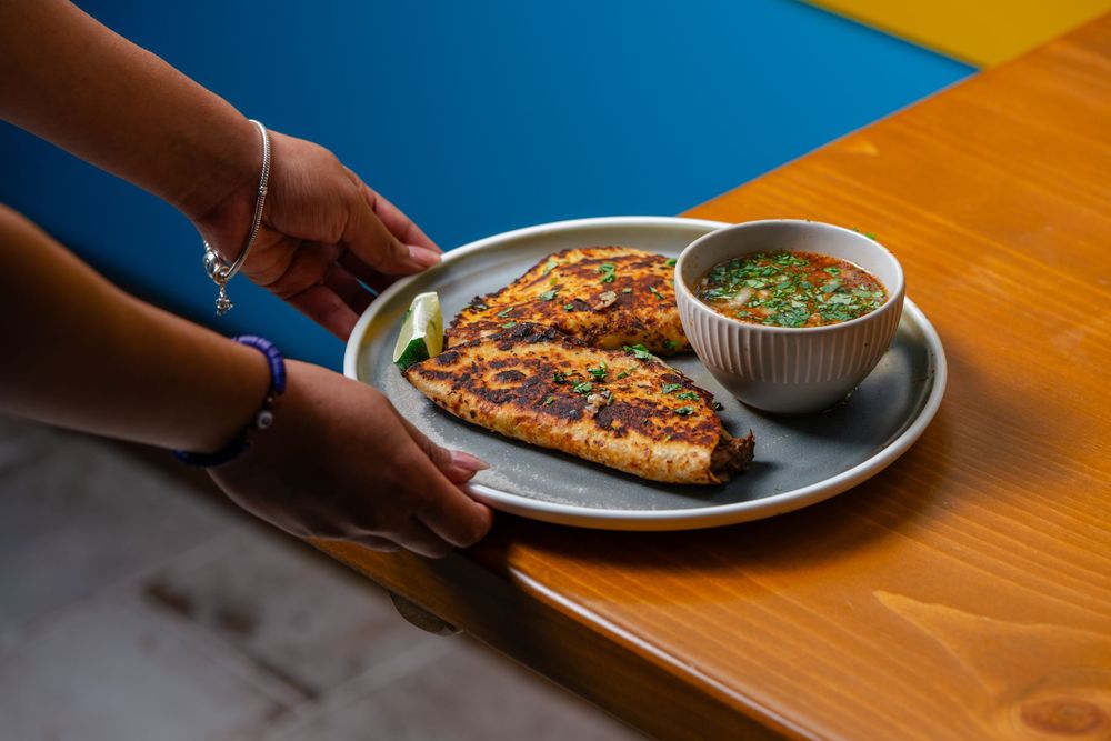 Hands serving a plate of grilled fish with fresh herb sauce in a small bowl on a wooden table.