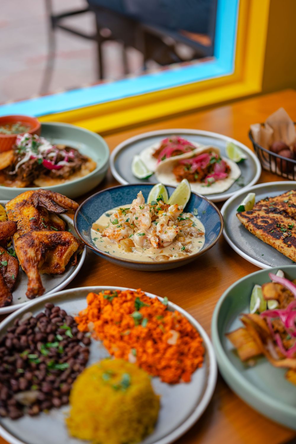 A vibrant spread of Latin dishes including shrimp, tacos, rice, beans, and roasted chicken on a wooden table.