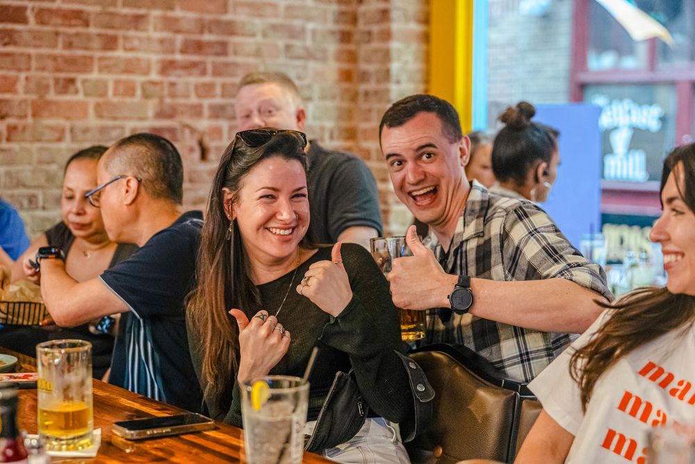 Friends smiling and giving thumbs up at a lively restaurant with brick walls and wooden tables.