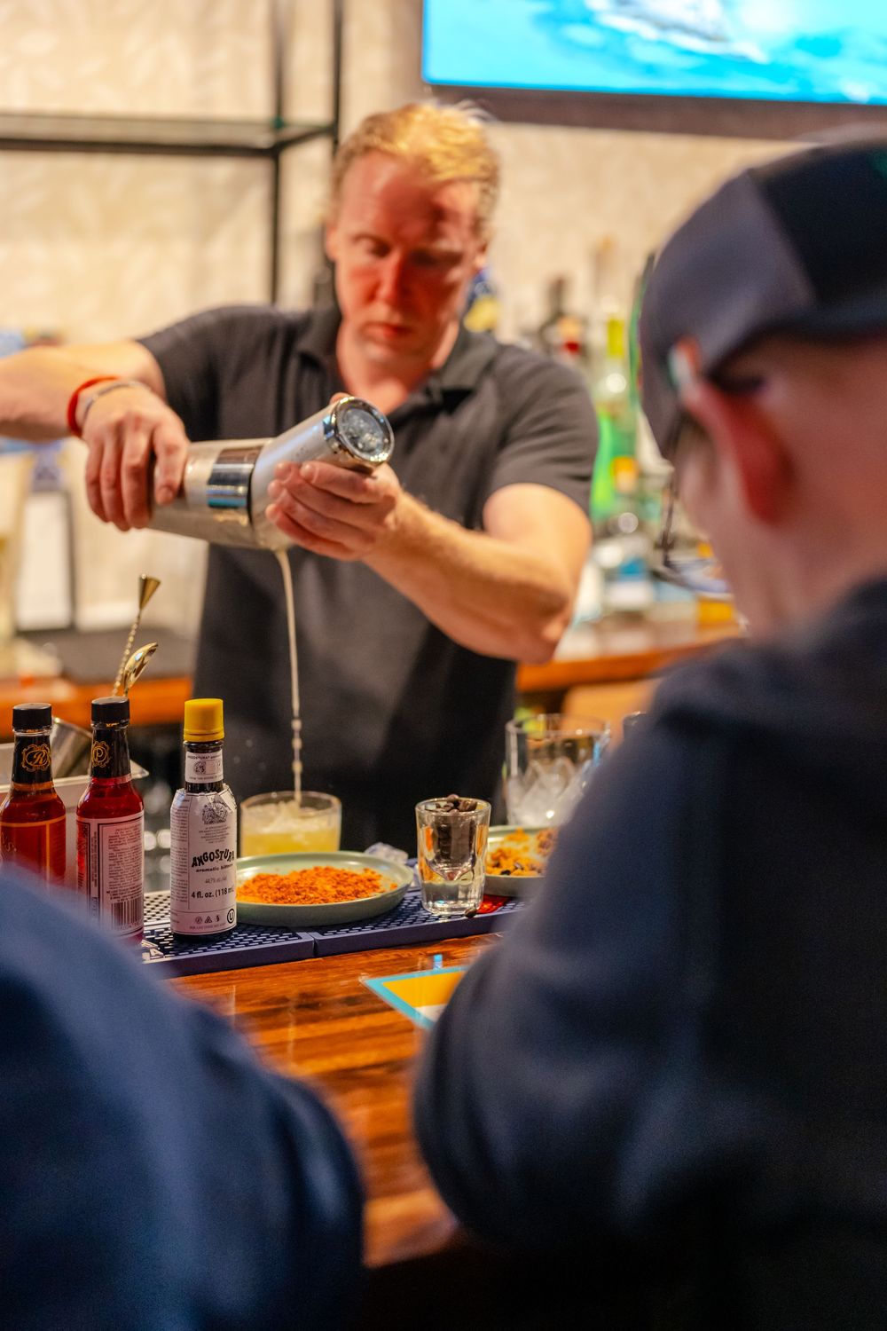 Bartender pouring cocktail shaker at bar counter with bottles and garnishes, serving customer in foreground.