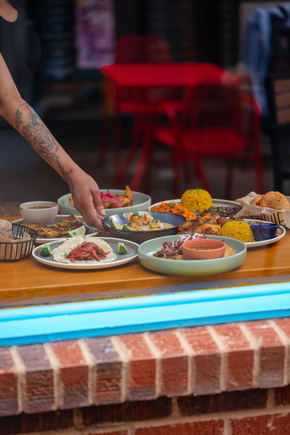 Tattooed hand arranging colorful dishes of food on a wooden counter with brick base at an outdoor restaurant.