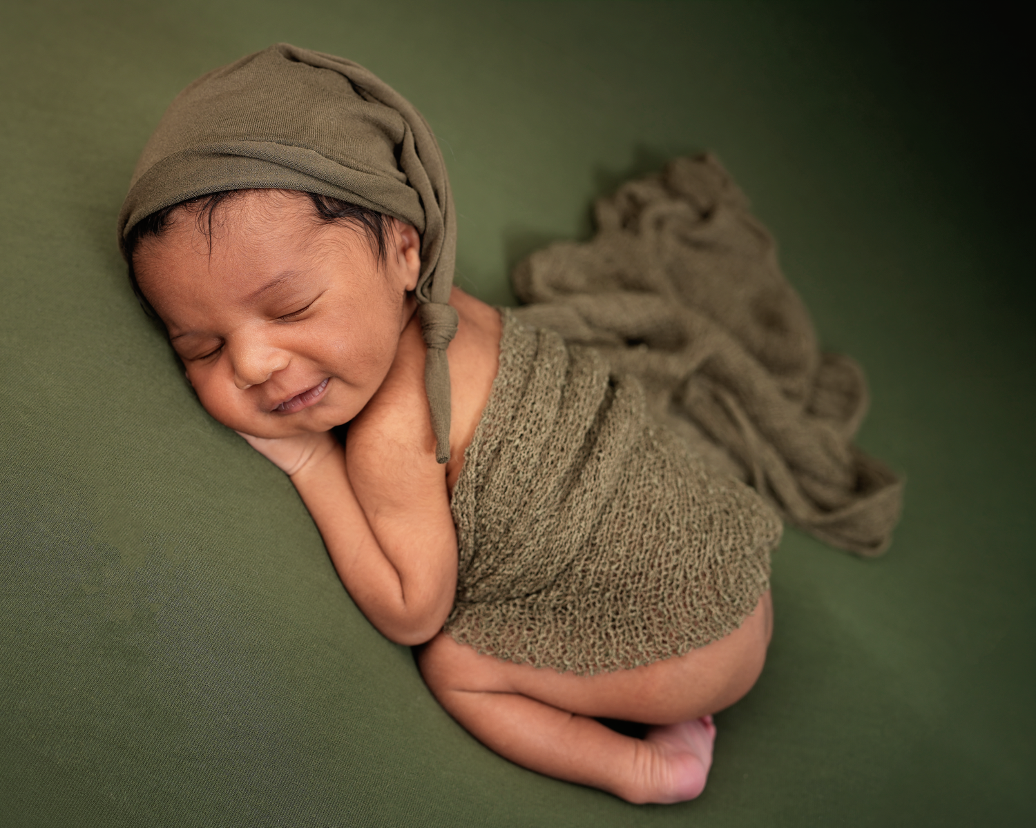Sleeping newborn baby smiling while comfortably wrapped in olive green during his photography session.